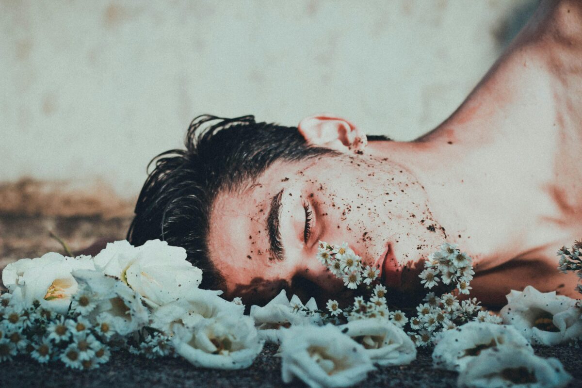 Young man is laying in a bed of beautiful white flowers and soil all over his face.