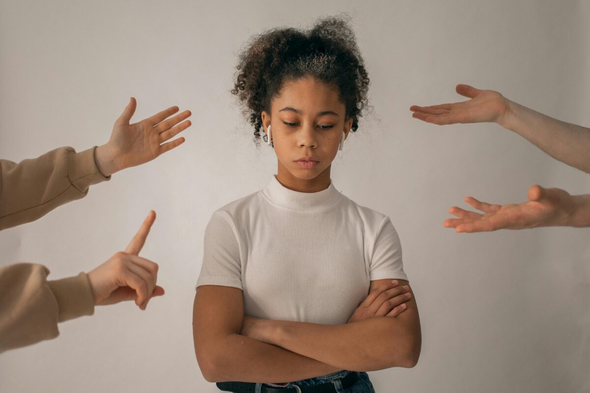 Young woman sad and frustrated with her arms crossed as her work colleagues point their fingers criticising her.