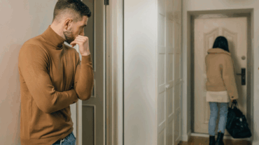 A man standing in the hallway at home watching his partner as she walks out the front door with her packed bag.
