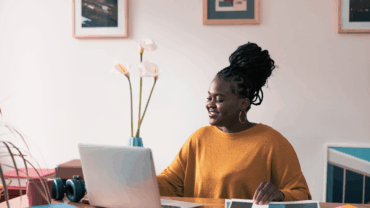 Happy young lady wearing a mustard yellow coloured sweater working on her laptop at home.