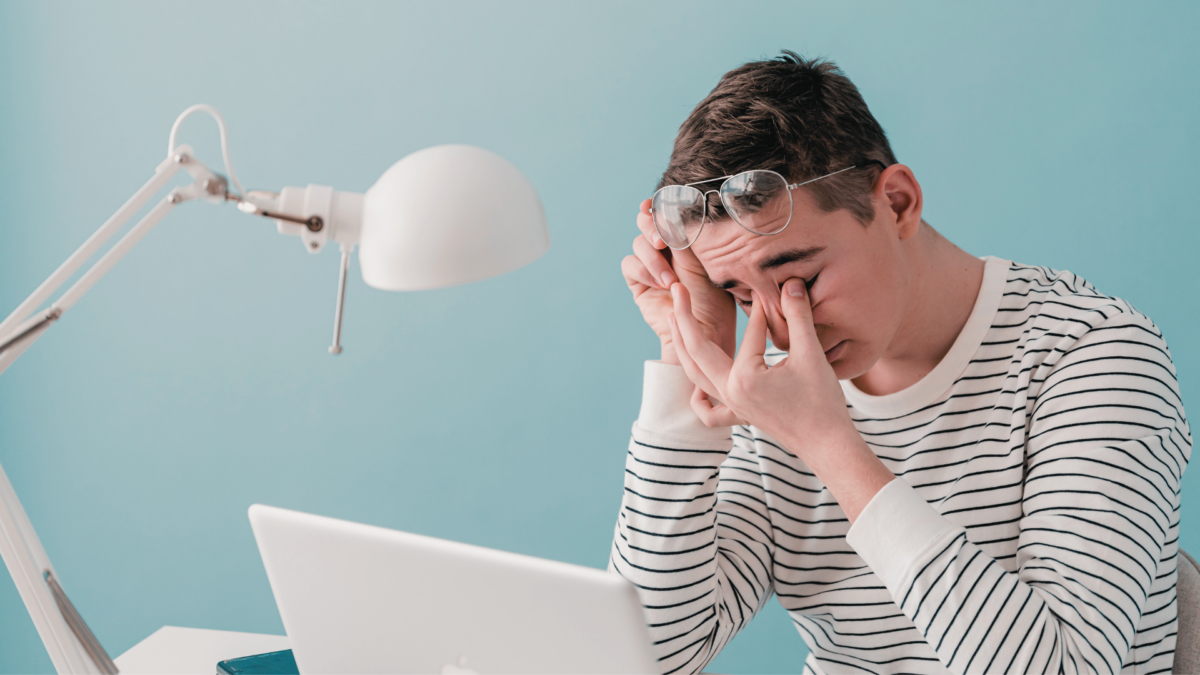 Man sitting at his desk pulls his glasses away from his eyes to rub them in exhaustion.