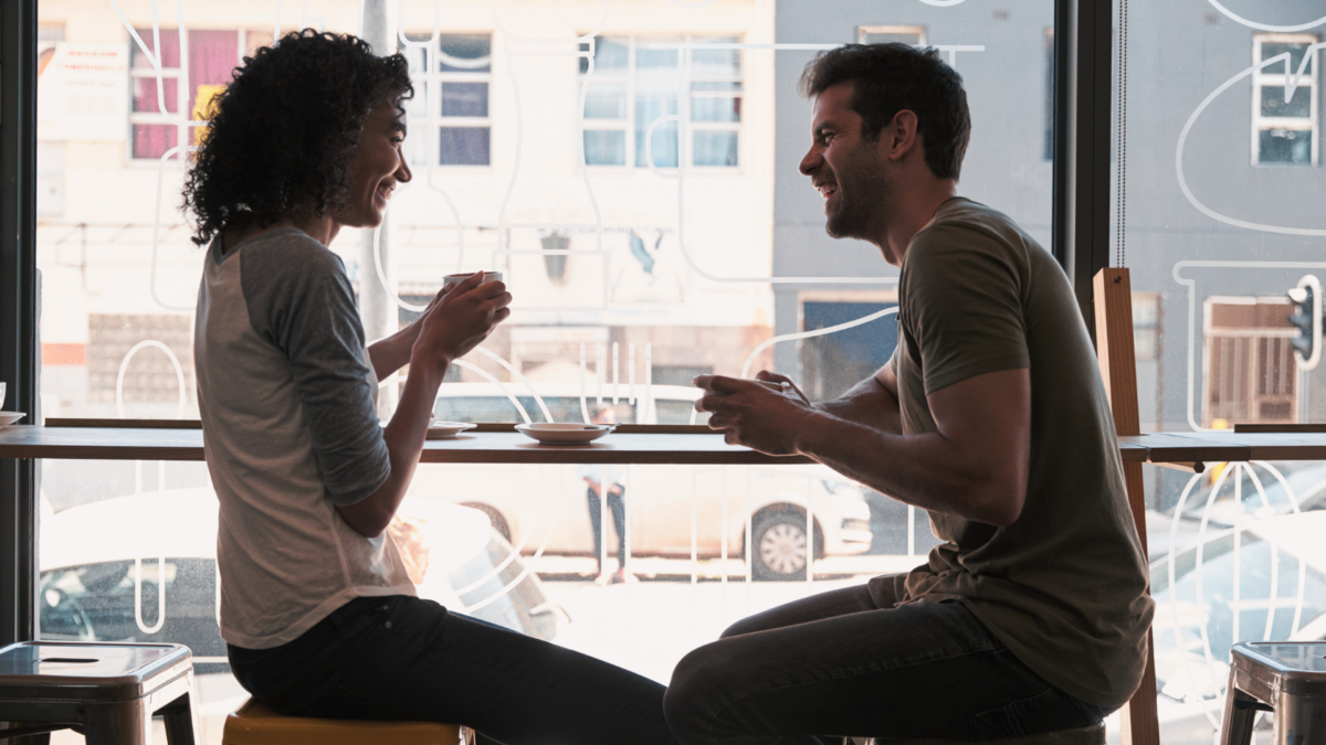 Man and a woman laughing and sitting by a large window, while on a date in an urban city coffee shop.