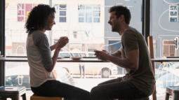 Man and a woman laughing and sitting by a large window, while on a date in an urban city coffee shop.